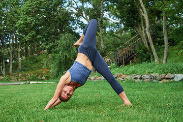 Obraz na płótnie Canvas athletic woman in blue sportswear practicing yoga while standing in an inverted pose on the grass outdoors