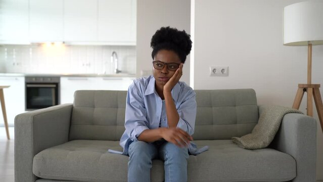 Flustered African American woman is very nervous sighing and touching face with hands sits on couch of home. Young black girl takes off glasses feeling fear after wrong decision or problems at work 