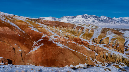 Fototapeta na wymiar The bright red-orange hillsides are covered with snow. A mountain range against the blue sky. Dry grass in the valley. Altai Mars Kyzyl Chin