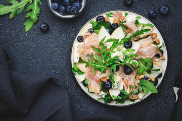 Tasty arugula salad with sweet pears, blueberries, roquefort cheese, smoked pork ham and crunchy walnuts. Black kitchen table background, top view, space for text