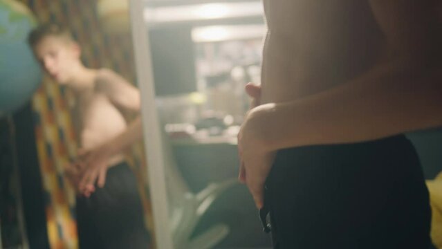 Determined young boy examines his abs and muscles in front of the mirror after exercise in his room. Bare upper body with muscular abs and shoulders. Weight training, fitness, physical activity.