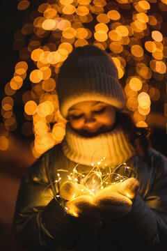 Girl and Christmas garland, selective focus. Smiling girl in warm clothes at night with blurry lights. Holidays theme with brown tint