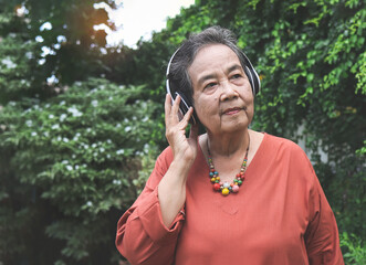 happy and healthy  Asian senior woman standing in the garden, listening to favorite music from headphones, smiling and looking away.
