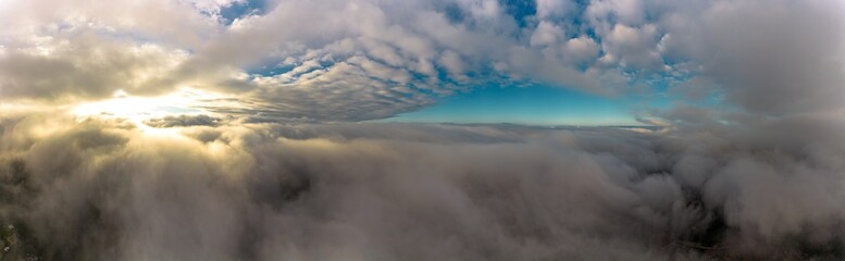 Aerial panorama view from between low clouds, illuminated by the sun, at sunrise. Captured using a drone.