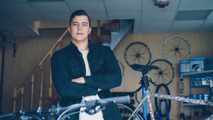 Portrait of handsome young man professional mechanic standing in bicycle repair workshop with his hands crossed and looking at camera. Bikes and shelves with spares are visible.