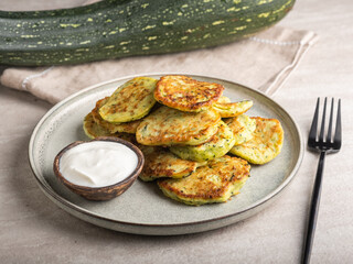 Vegetable vegetarian zucchini pancakes with sour cream on a round plate and green zucchini background, summer food, delicious snacks.