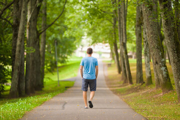 Young adult man slowly walking through alley of green trees on sidewalk at city park in warm summer...