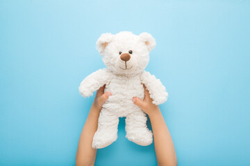 Baby boy hands holding smiling white teddy bear on light blue table background. Pastel color....