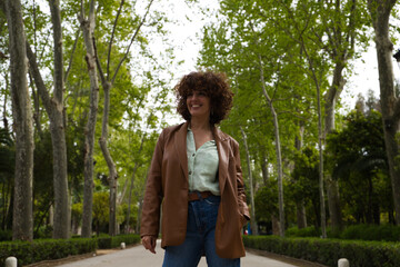 A middle-aged, beautiful, curly-haired Spanish woman stands in a park with large trees in a European city. The woman is dressed in modern and trendy clothes. Travel and holiday concept.