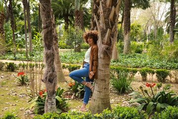 Naklejka na ściany i meble A middle-aged, beautiful, curly-haired Spanish woman stands in a park with large trees in a European city. The woman is dressed in modern and trendy clothes. Travel and holiday concept.