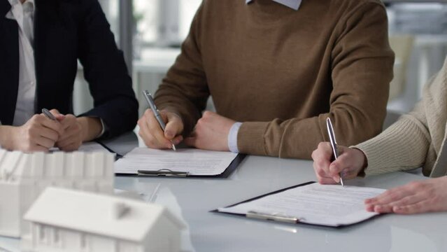 Panning hands shot of man and woman sitting at table with scaled house models in real estate agency and each putting signature on separate copies of legal documents, and professional lady waiting