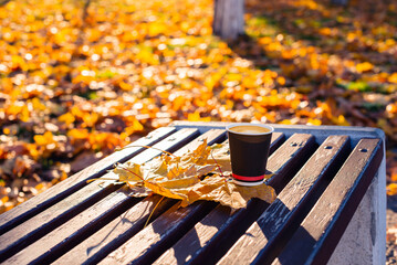 A cup of coffee stands on a bench with autumn leaves. 