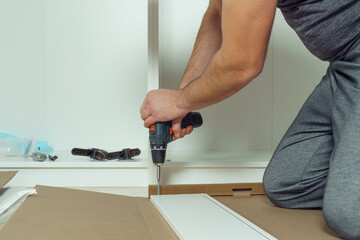 Male hands hold cordless electric screwdriver, screw to fix walls of white drawer, assembling dresser or wardrobe on floor. Self-assembly of furniture at home, DIY, furniture repair and rearrangement