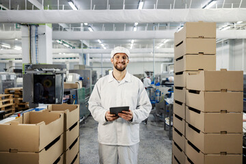 A food factory supervisor stands between the boxes with meat, holds a tablet, and smiles at the...