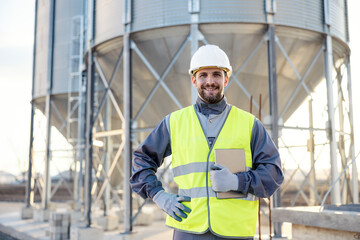 Naklejka na ściany i meble A supplies supervisor proudly stands in front of the silos, with a tablet in his hands, and smiles at the camera.