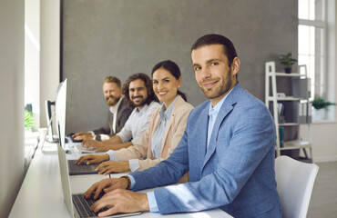 Team of happy young business people working on modern computers. Group portrait of successful cheerful coworkers and teammates sitting in row at office desk with laptops, looking at camera and smiling