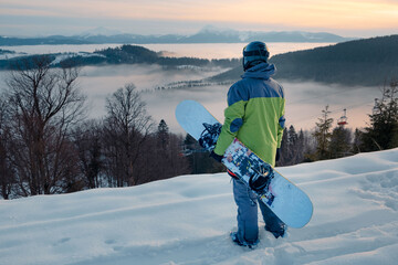 man snowboarder enjoying the view of sunset above the snowed mountains