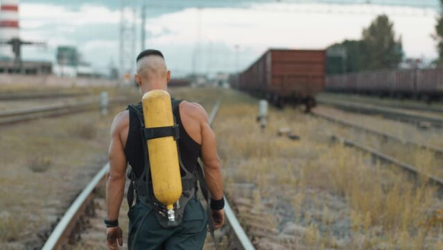 Young muscular man goes along the rails of marshalling yard and removes a drager compressed air tank from his back