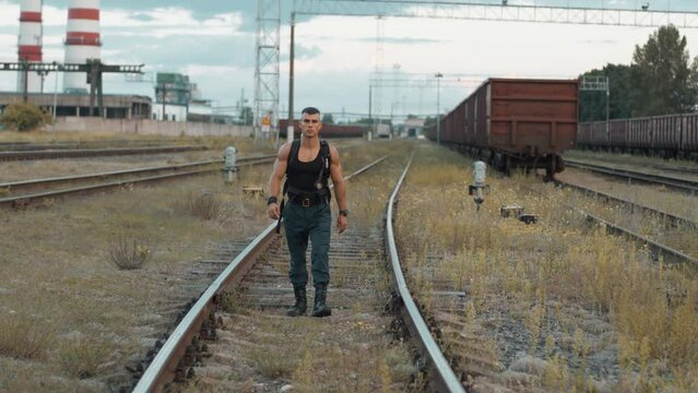 Young muscular man with balloon dragger on the back goes between the rails at railway yard near factory. Street workout