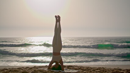 Fototapeta na wymiar Woman doing headstand yoga pose on seashore at sunrise. Girl training balance.