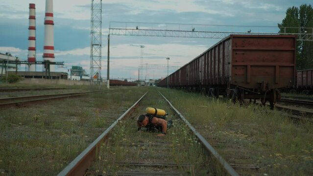 Young muscular man with balloon drager on his back training at railway yard near the factory and moves between the rails doing push-ups