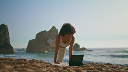 Woman watching yoga online lesson using modern tablet on sand beach morning time
