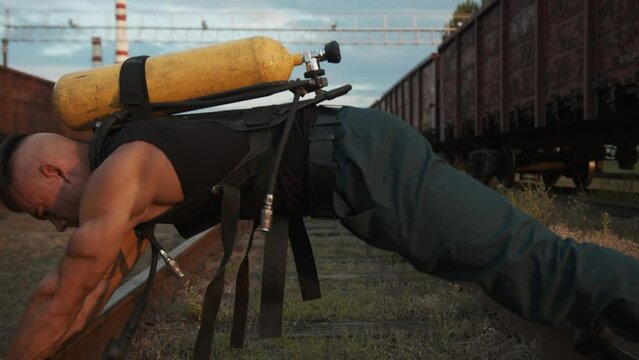 Bodybuilder training at the railway yard and moves along the rails in the exercise plank with drager compressed air tank on his back