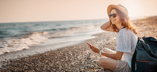 Portrait of young cheerful woman in a hat and sunglasses sitting with a backpack on the seashore at sunset