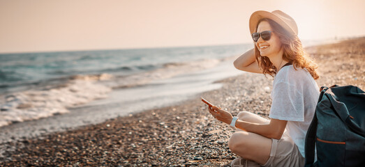 Portrait of young cheerful woman in a hat and sunglasses sitting with a backpack on the seashore at...