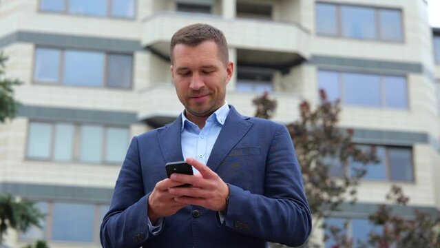 Happy handsome confident guy in suit typing on mobile phone and smiling in good mood. Close up of joyful businessman texting message on smartphone. Slow motion. Urban concept