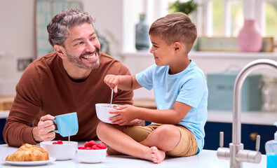 Family With Father In Kitchen With Son Sitting On Counter Eating Breakfast Together
