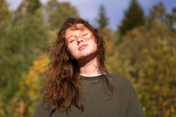 Portrait of happy relaxed young beautiful woman, calm girl is relaxing, meditating breathing deep deeply fresh autumn air at sunny day in forest with eyes closed, smile. Sunshine, natural background