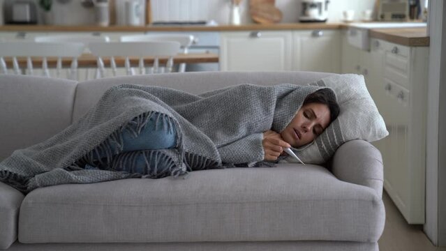 Sick European woman millenial with flu sneezes and measures temperature with thermometer lying on sofa in living room of apartment. Caucasian lady coughs and suffers from runny nose due to cold snap
