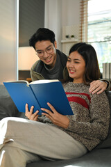 Smiling young couple relaxing on couch and reading book, spending leisure time on winter holidays at home