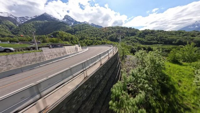 FPV sport drone shot over cable way empty serpentine asphalt road mountain alpine resort sunny landscape aerial view. Speed flight in bridge tunnel picturesque suburb city landmark infrastructure 4k