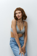 vertical studio photo of a beautiful, tanned woman with a toned, athletic body, posing relaxed against a light background while standing in stylish summer clothes leaning on the wall