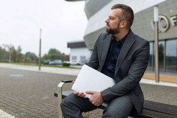 portrait of successful mature businessman with laptop sitting next to office building