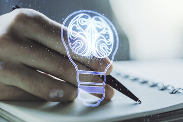 Double exposure of creative light bulb hologram with human brain and with man hand writing in notebook on background, idea and brainstorming concept