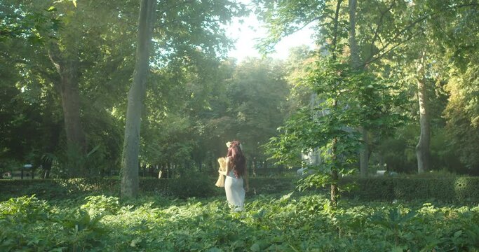 Beautiful long fashion shot of a woman dressed as a goddess with a white dress and a bouquet of roses walking in a forest with the sunlight hitting it in the morning time.