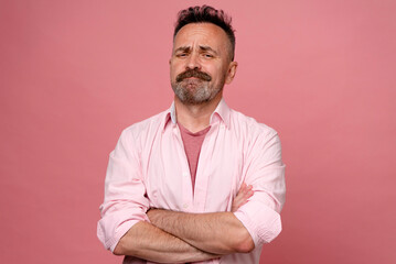 Portrait of doubtful man in pastel pink shirt on pink wall. Middle age grey-haired beard man with crossed hands thinking about question, pensive expression. smiling with thoughtful face. doubt concept - obrazy, fototapety, plakaty