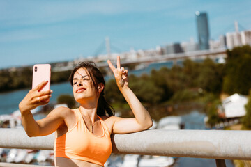 Happy beautiful woman in sportswear taking selfie with her smartphone on the bridge. Added grain film look effect