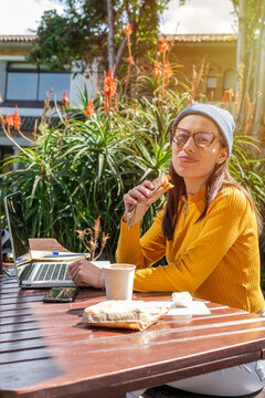 Hispanic female student sitting on college campus in front of her computer and notebook taking a break while eating a snack