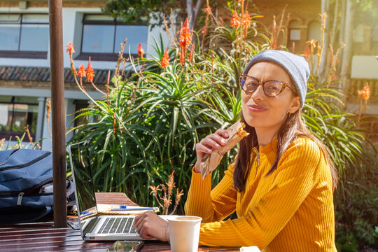 Hispanic Female Student Sitting On College Campus Taking A Break In Front Of Her Laptop While Eating A Snack