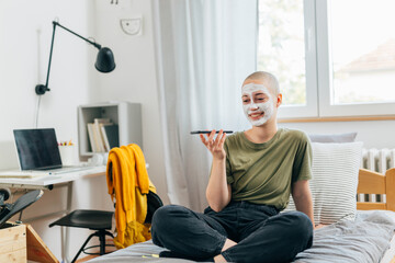 shave head teenager female with applied cosmetic face mask talking on mobile phone in her room