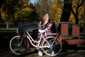 Obraz premium The girl is sitting on a bench in the autumn park. Eats ice cream. Dressed in autumn clothes. There is a bicycle nearby. Autumn city park.