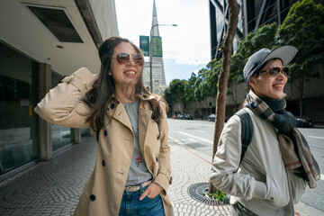 Naklejka premium happy asian businesswoman holding her messy hair while walking with her colleague on street during lunch break on windy day in san Francisco with Transamerica Pyramid at background