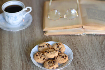 Cup of tea, plate with chocolate chip cookies, open book, reading glasses, lit candles and dry lavender flowers. Hygge at home, selective focus.