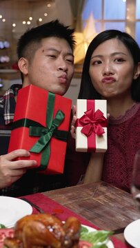 Vertical Screen: cheerful asian couple making funny faces while taking selfie photos with their Christmas presents using mobile phone at xmas eve dinner in dining room