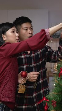 Vertical Screen: happy Japanese asian husband and wife working together to decorate the Christmas tree with hanging ornaments while preparing for the coming winter holiday at home