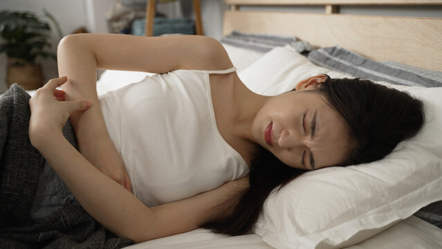 asian girl having severe cramp in lower abdomen is holding her belly and lying on bed with a groan of pain in the bedroom at home during daytime.